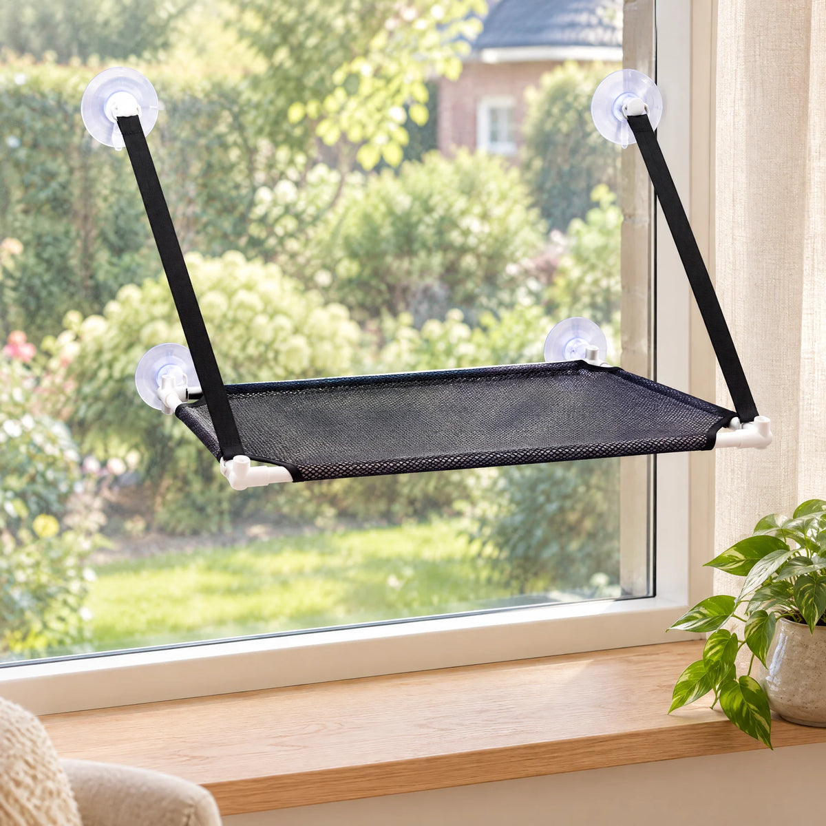 Zwarte kattenhangmat hangt met vier zuignappen en twee banden aan het raam van een woonkamer, met uitzicht op de tuin