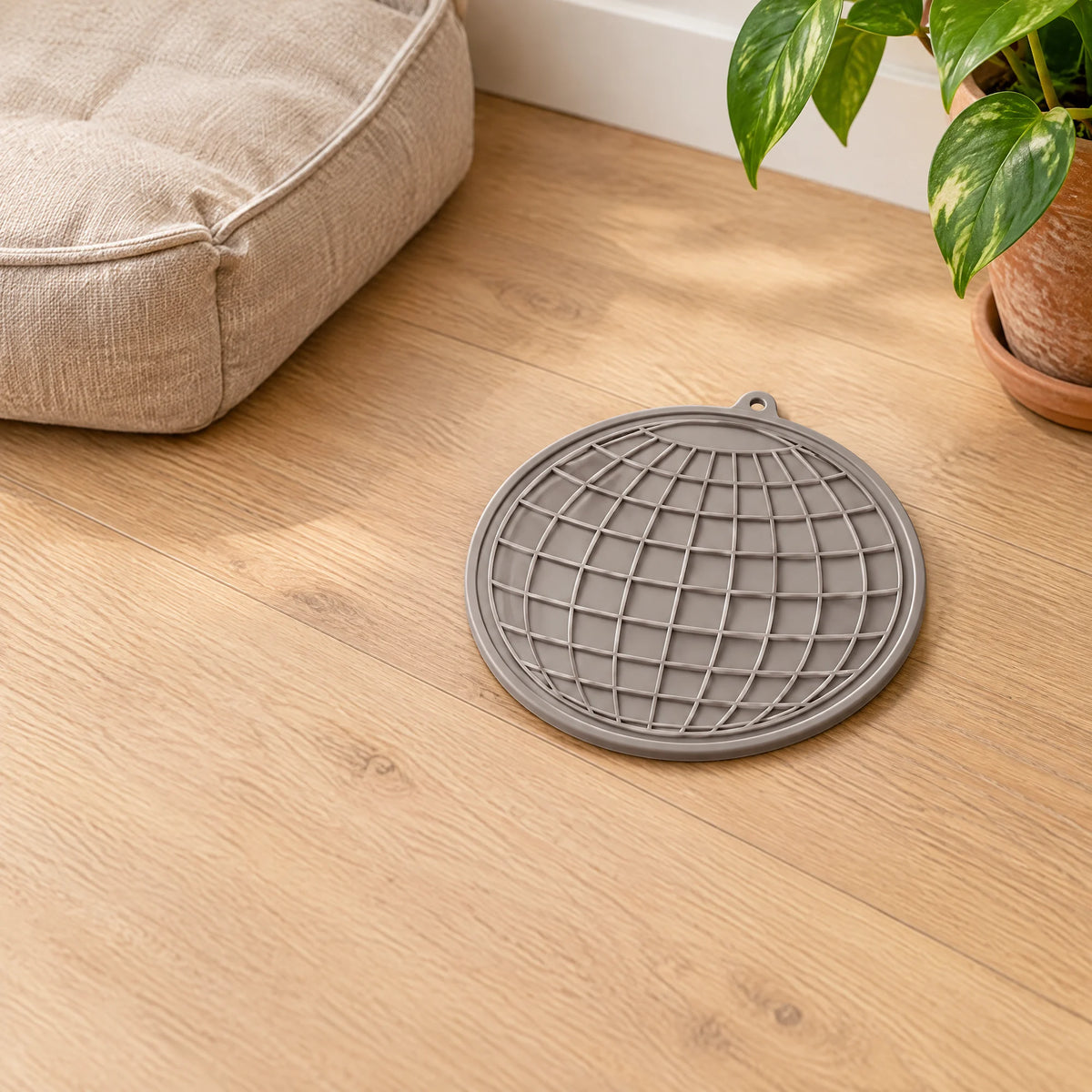 Ronde grijze siliconen likmat voor hond en kat op een eiken vloer naast een hondenkussen