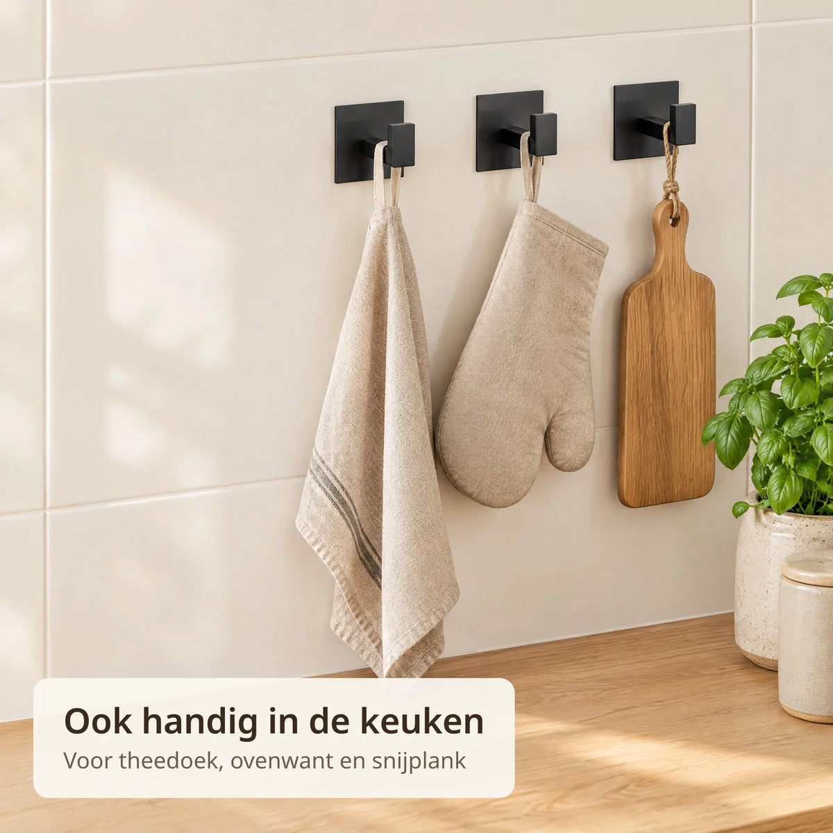 Drie zwarte zelfklevende haken op keukentegels met een theedoek, een ovenwant en een houten snijplankje